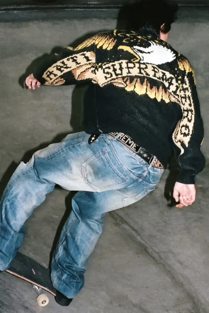 A skateboarder carves along a concrete surface wearing a black knit Supreme × Antihero sweater featuring a large woven eagle graphic across the back, paired with faded baggy jeans, captured in motion from behind