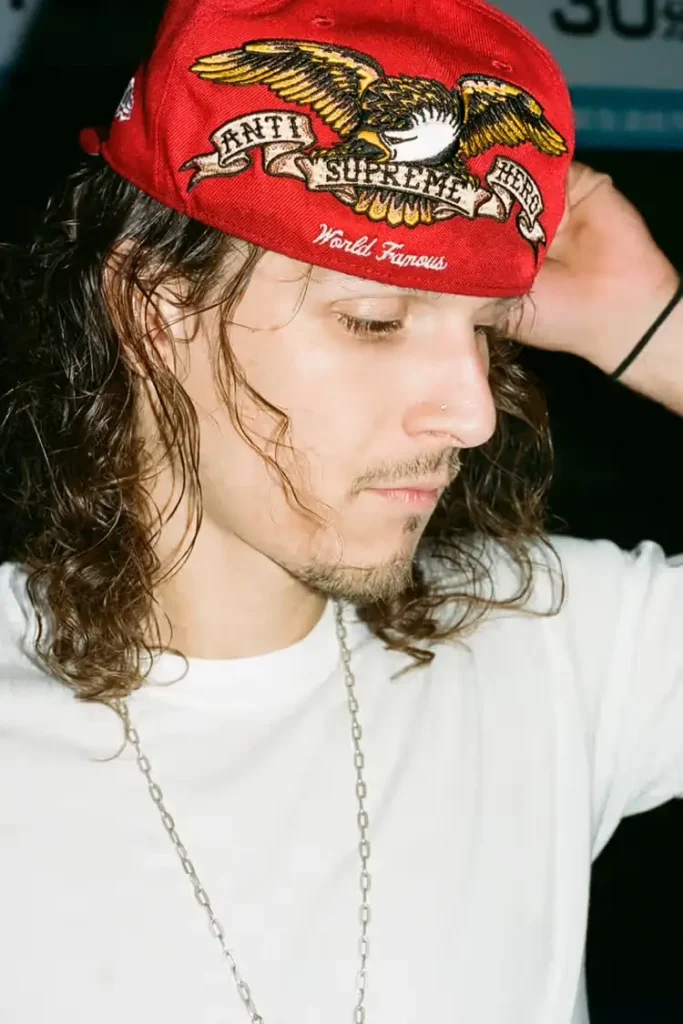 A person with long wavy hair wears a red Supreme × Antihero cap featuring a large embroidered eagle graphic and “World Famous” lettering, shown in a close-up portrait against a dark background