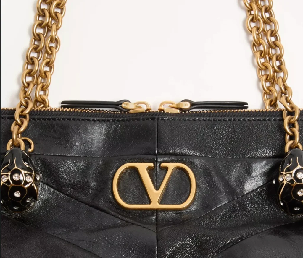 Detail view of the Valentino Garavani Panthea Shoulder Bag highlighting the central gold VLogo emblem on black nappa leather with chevron stitching. The image showcases the antique gold chain handles and black enamel feline-head charms adorned with crystal embellishments, emphasizing Valentino’s refined craftsmanship and haute hardware detailing