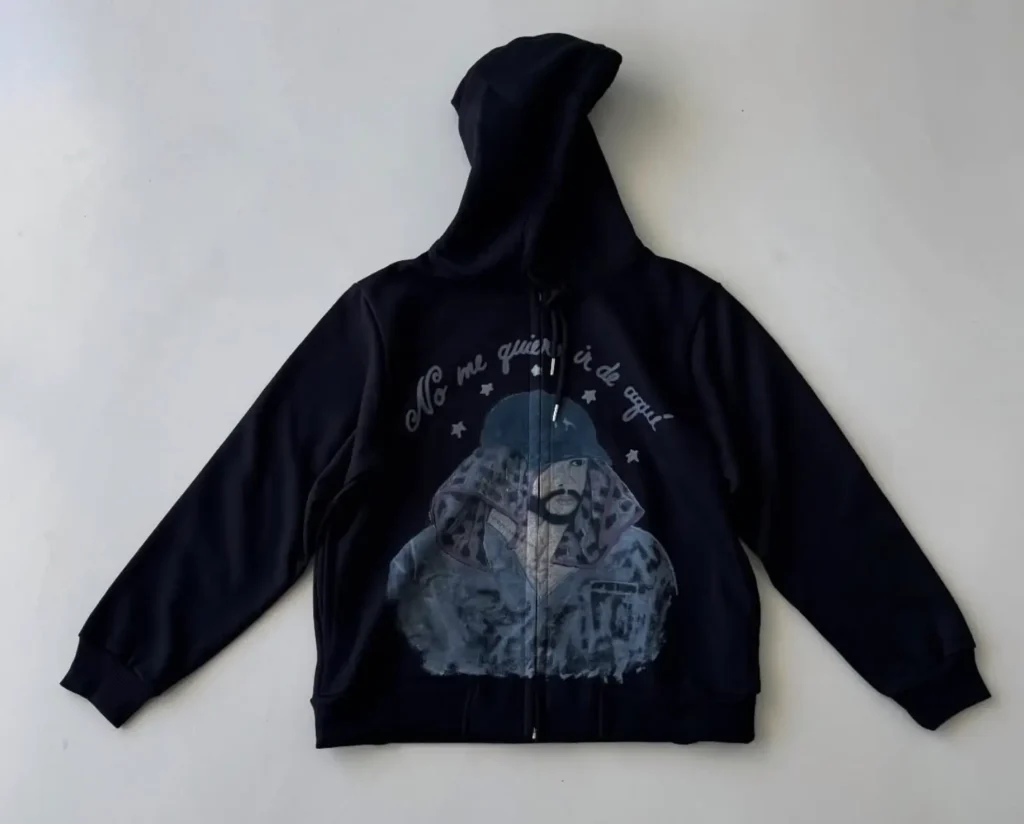 Black full-zip hoodie featuring an illustrated portrait of Bad Bunny wearing a hood and patterned scarf, with the phrase “No me quiero ir de aquí” curved above in stylized lettering and stars — part of the official Bad Bunny × FRSH Company Puerto Rico residency merchandise collection