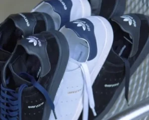 A close-up angled shot of several pairs of Adidas Stan Smith sneakers from the “everyone” collection, stacked together in a casual arrangement. The group includes black suede pairs with tonal black details and navy-striped tongues, and light grey pairs with white laces and navy heel tabs. The shoes rest against a metal mesh surface, with the Adidas trefoil logos and “everyone” branding clearly visible on the tongues and side panels