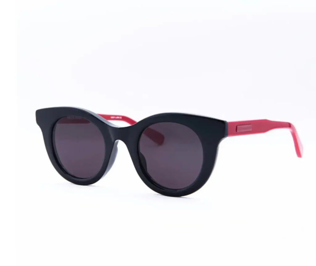 Black rounded cat-eye sunglasses with matte dark lenses, featuring bold pink temples and a clean, angled three-quarter view against a bright white background