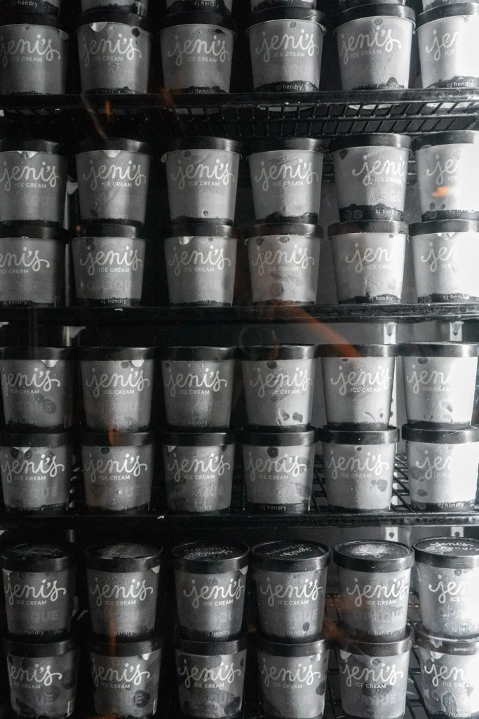 Rows of Jeni’s Splendid Ice Cream pints stacked in a freezer, featuring sleek matte-black lids and minimalist silver “Jeni’s x CJ Hendry” branding, emphasizing the monochrome design of the collaboration’s limited-edition packaging