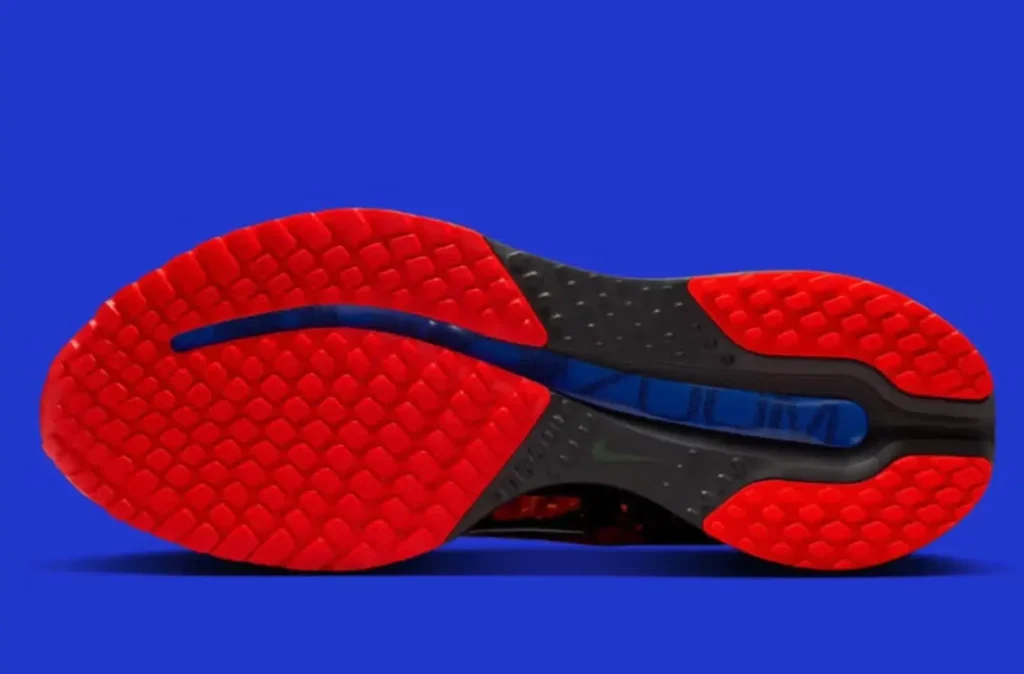 Bottom view of the Nike Pegasus Premium “Bright Crimson” outsole, showing the red waffle-pattern traction zones, black midfoot support, and a blue flex groove running through the center against a vivid blue background.