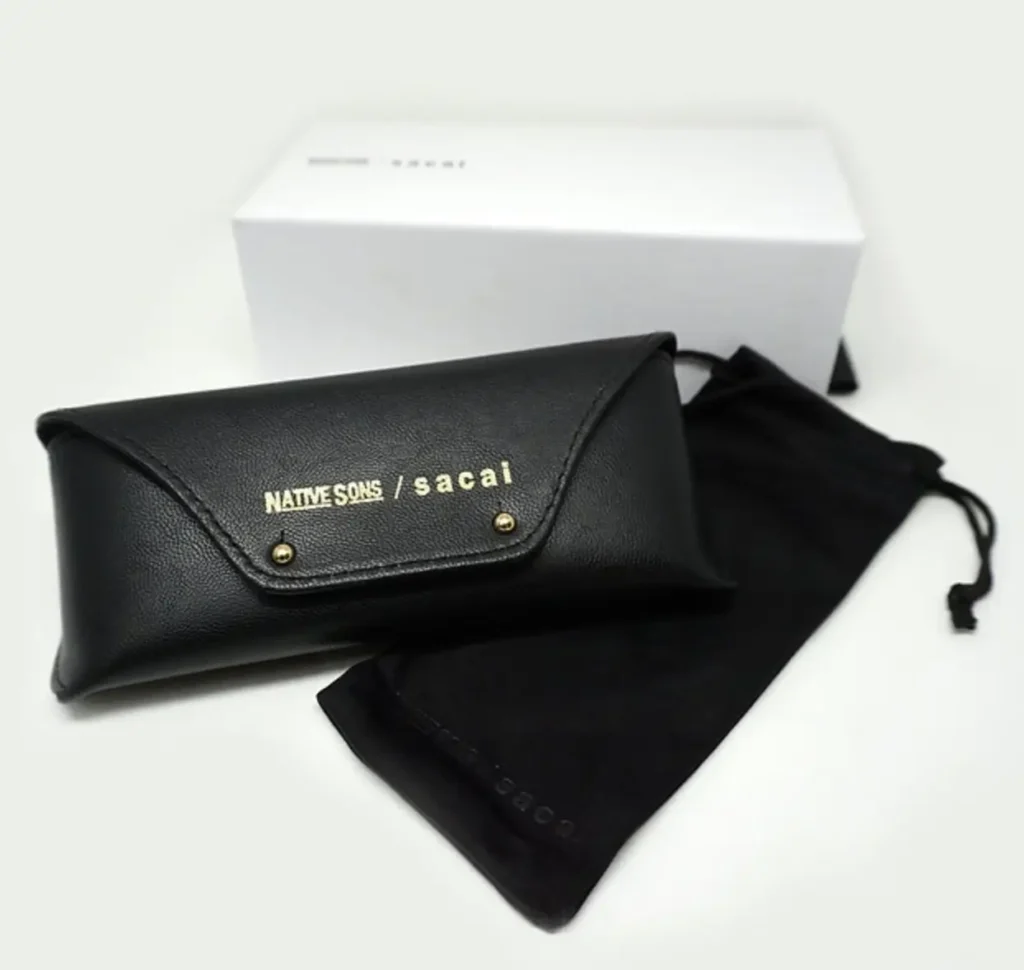 Black leather eyewear case with gold “NATIVE SONS / sacai” branding on the front flap, shown alongside a black drawstring pouch and a white branded box, all arranged on a clean light background