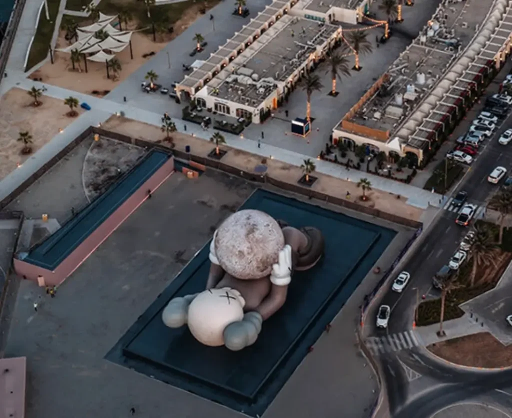 An aerial view of the massive KAWS Companion sculpture in Abu Dhabi, shown reclining on a dark platform while holding a large moon-like sphere. Surrounding it are illuminated palm-lined walkways, nearby buildings, parked cars, and curving roads, all captured at dusk with soft, muted lighting