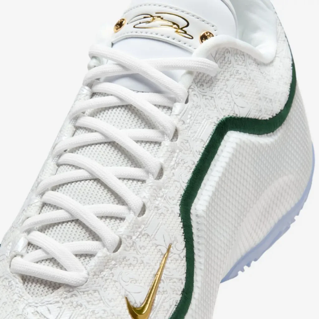 Close-up angled view of the Nike LeBron 23 “From This Point Forward,” highlighting the white textured upper with intricate embossed patterns, green embroidered trim, and a metallic gold mini Swoosh near the forefoot. White laces and a smooth white leather tongue with LeBron’s gold signature complete the premium look