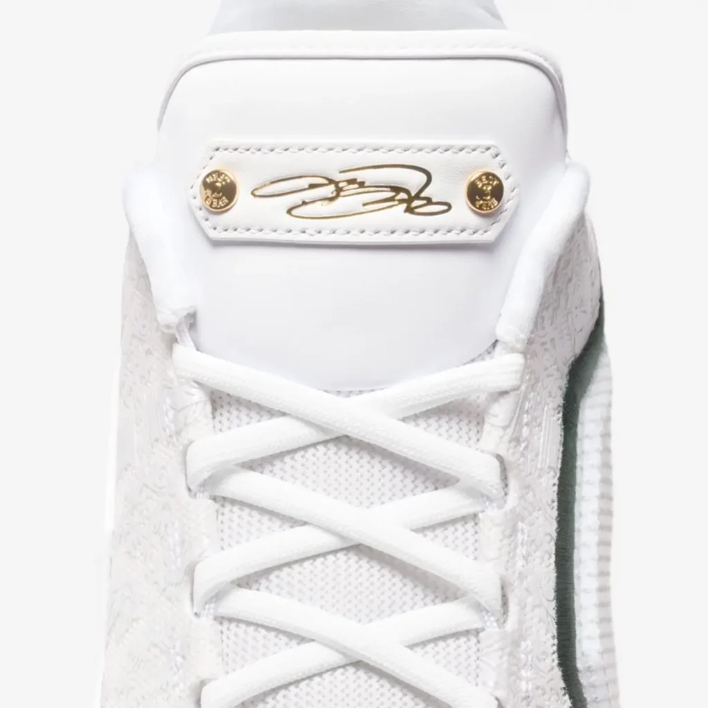 Close-up of the Nike LeBron 23 “From This Point Forward” tongue, showcasing a white leather patch with LeBron James’ gold signature and two gold rivets, above white laces and a textured white upper with green trim along the side