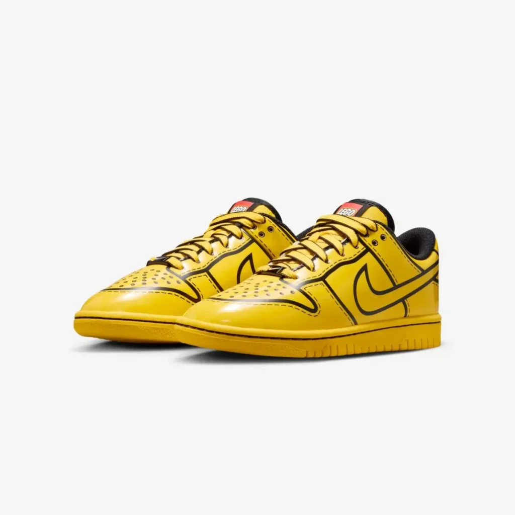 A pair of LEGO x Kids Nike Dunk Low “Tour Yellow” sneakers displayed at a three-quarter angle, featuring bright yellow patent-like leather with bold black outline detailing, a matching yellow sole, black inner lining, and a red LEGO logo tab on the tongue. The shoes sit against a plain white background