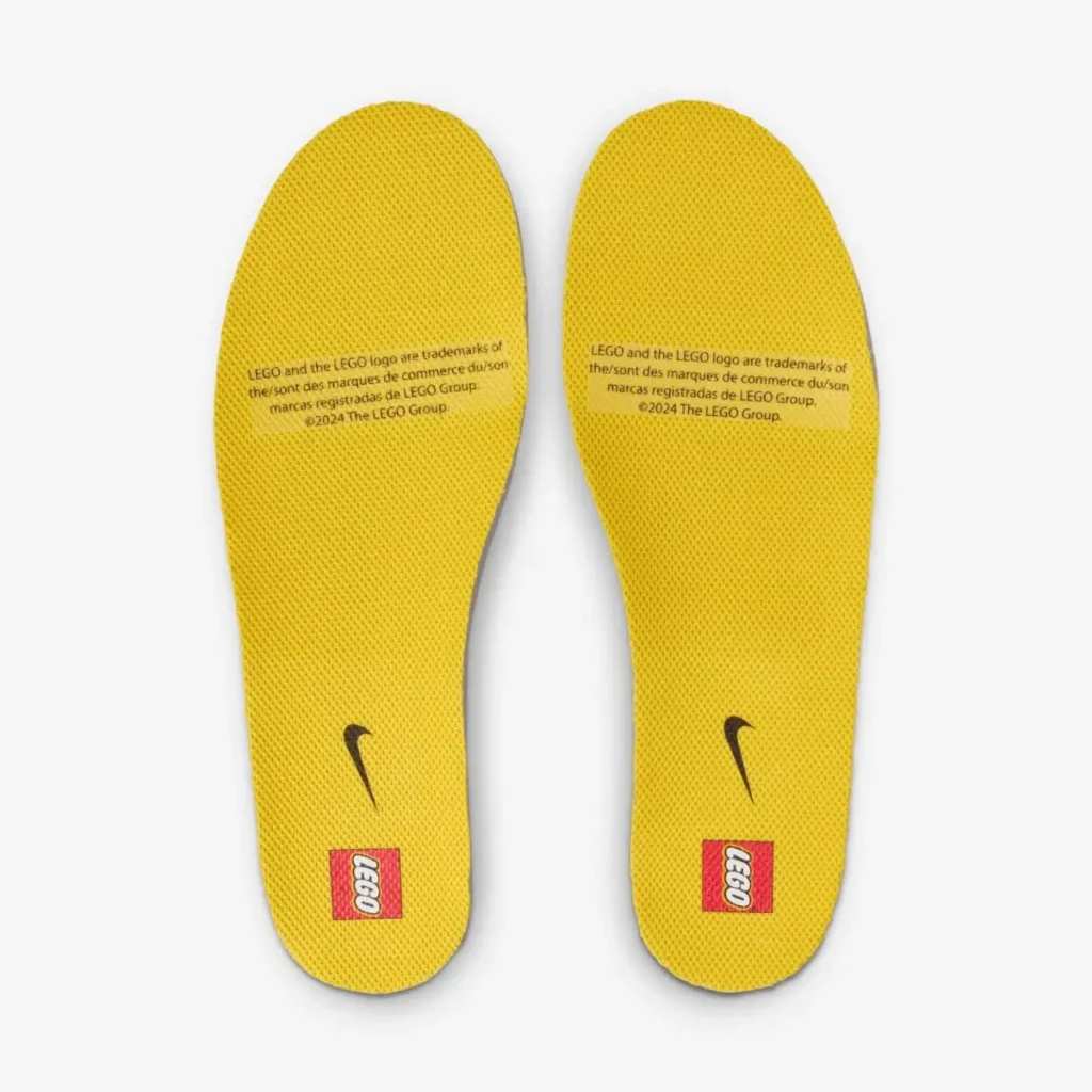 Top-down view of the LEGO x Kids Nike Dunk Low “Tour Yellow” insoles in bright yellow, featuring a textured surface, small black Nike Swoosh near the arch, and red LEGO logo blocks at the heel. Both insoles display printed LEGO trademark text, set against a plain white background