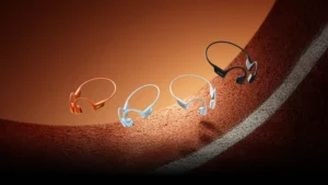 Four Shokz OpenRun Pro 2 bone-conduction sports headphones in orange, sky blue, white, and black appear suspended above a curved running-track surface under warm studio lighting, symbolizing speed, endurance, and motion.