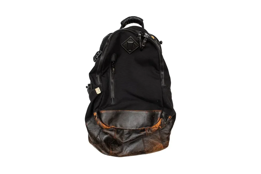 A visvim backpack in olive technical fabric featuring utility zippers and leather pull tabs, finished with a quilted patchwork bottom panel made from mixed geometric fabrics in cream, brown, and plaid tones