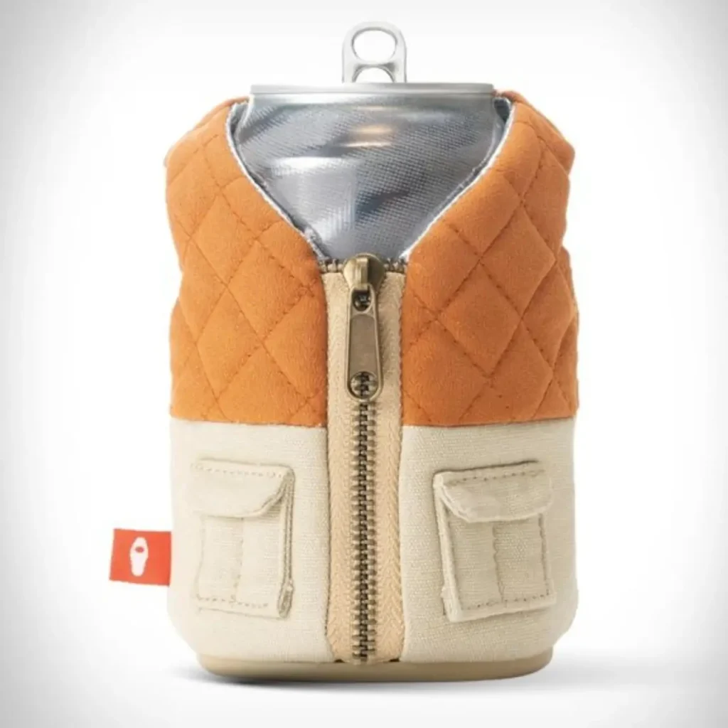 A beverage can wrapped in a Puffin Drinkware holder designed like a quilted vest, featuring an orange diamond-stitched top, cream canvas lower panels with flap pockets, and a central brass zipper that mimics rugged outdoor apparel