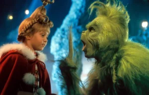 Still Scene from the Grinch movie, left Taylor Momsen young child dressed in a red, fur-trimmed holiday cloak looks up calmly at a green costumed figured Jim Carrie, a furry creature with exaggerated facial features who is raising a finger while speaking. The scene is brightly lit with a whimsical, snowy background, capturing a moment from a fantasy Christmas film