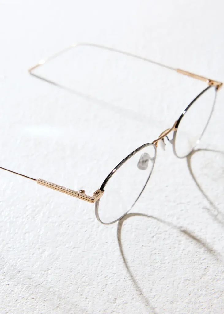 A close-up of round eyeglasses with thin silver rims and delicately engraved gold temples, resting on a textured white surface with soft shadows cast from the frame