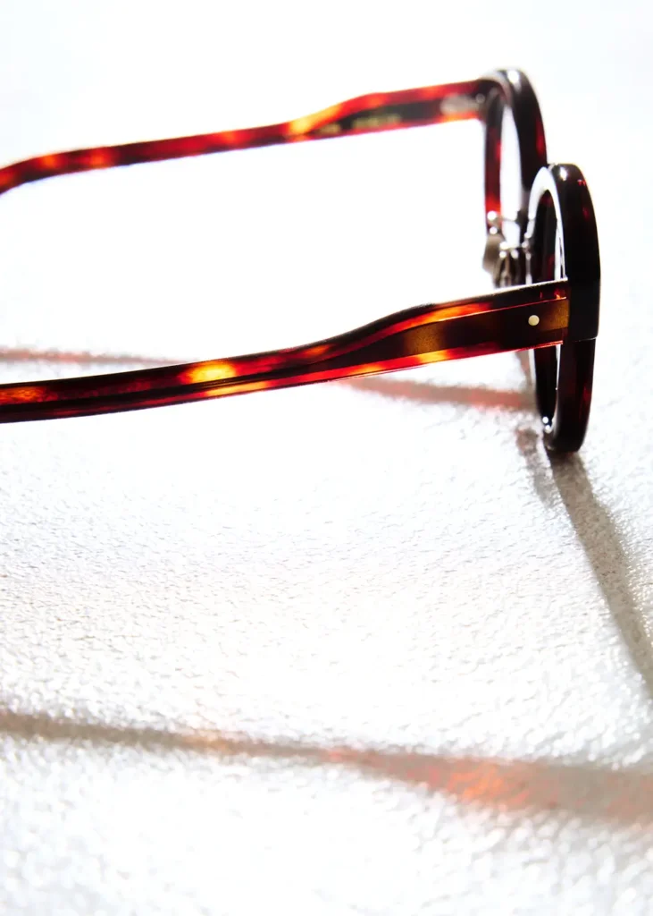 A close-up of tortoiseshell eyeglass temples glowing with warm amber highlights as light passes through the acetate, casting soft shadows across a textured white surface