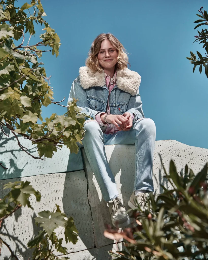 Person wearing a light blue Levi’s sherpa-collared denim jacket and matching jeans, sitting outdoors on a concrete ledge with greenery in the foreground and a clear blue sky behind them