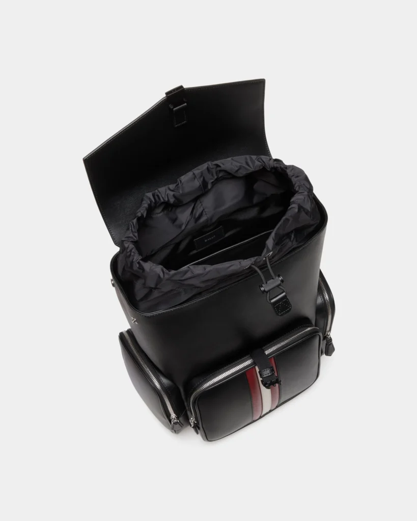 Top-down view of the Bally Mythos Backpack in black recycled leather, showing the structured flap lifted to reveal the drawstring opening, spacious black interior, and organized internal compartments alongside the front pocket with red and cream stripes