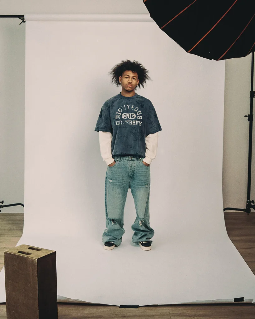 Model wearing the WE ARE RIGHTEOUS ‘Righteous University’ Thermal L/S, styled with distressed baggy denim and shoes, standing in a studio setup with seamless backdrop and overhead lighting