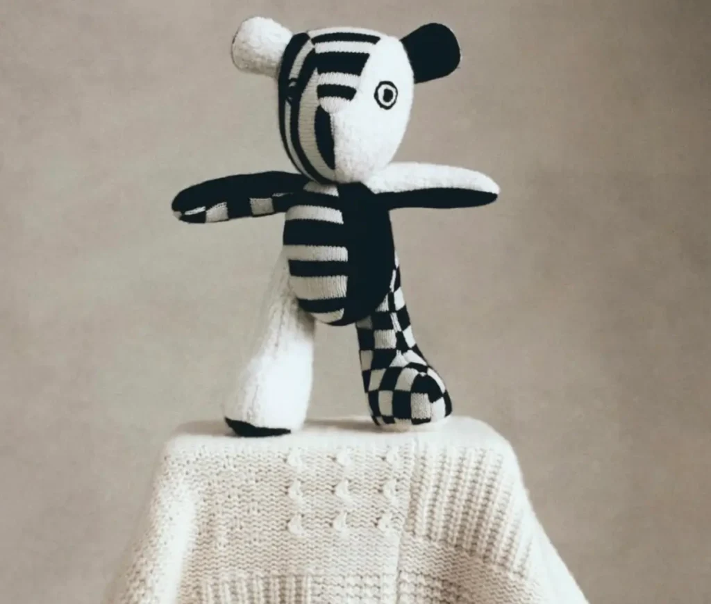 The Elder Statesman Cashmere Teddy in black-and-ivory striped and checkerboard knit, posed playfully on a textured cream cashmere blanket against a soft beige backdrop
