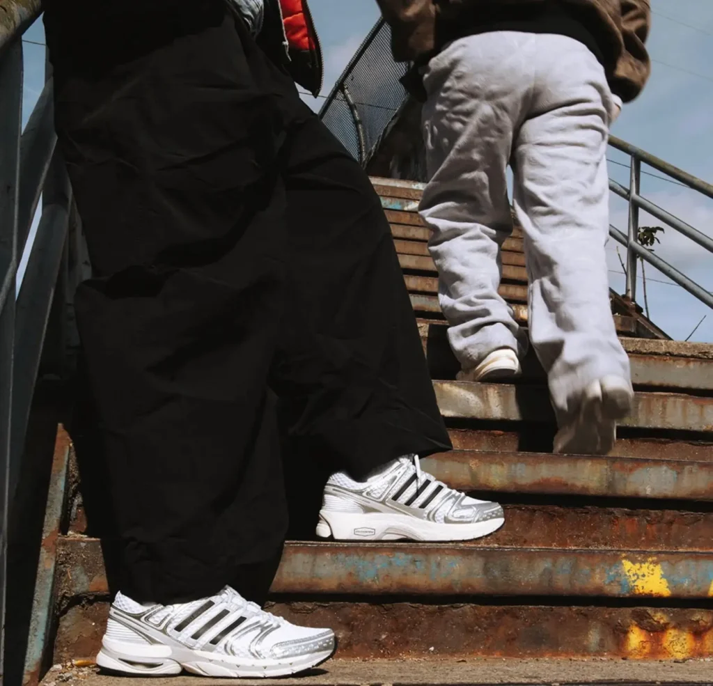 A close-up of two people walking up rusted outdoor stairs, showcasing retro-tech adidas Adistar sneakers in white with black stripes paired with oversized black pants and grey sweatpants, set against an urban industrial backdrop