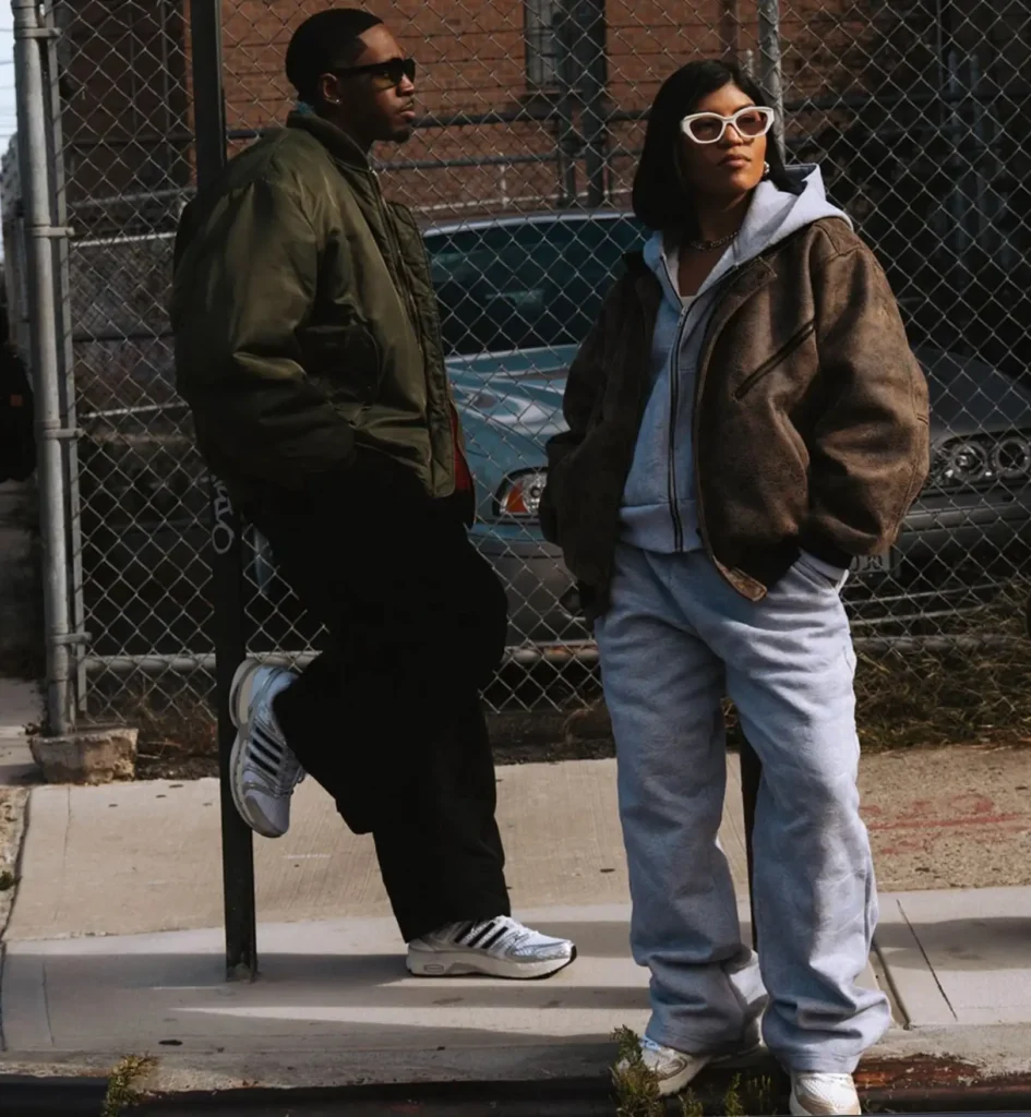 Two people wearing oversized streetwear jackets and sweatpants stand on a sidewalk in front of a chain-link fence, both styled in retro-tech adidas Adistar Control sneakers. The scene has an urban, industrial feel with muted tones and a parked car in the background