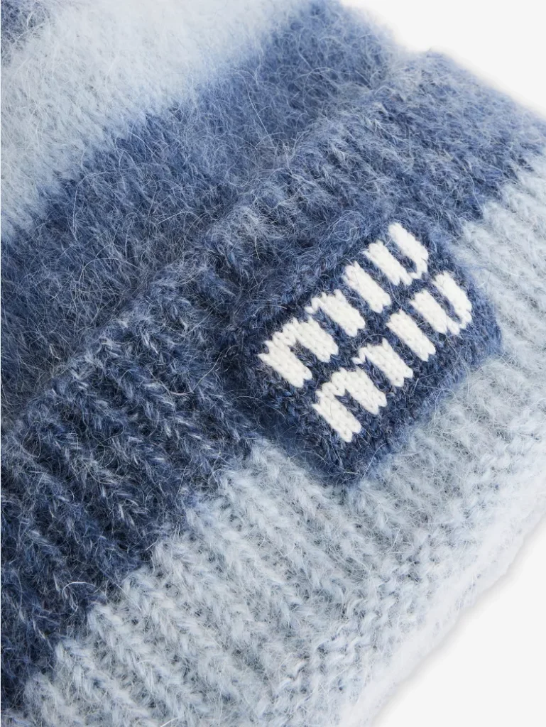 Close-up detail of the Miu Miu striped wool-blend beanie, highlighting the soft fuzzy knit texture, blue and light-blue stripes, and the embroidered Miu Miu logo patch on the ribbed cuff