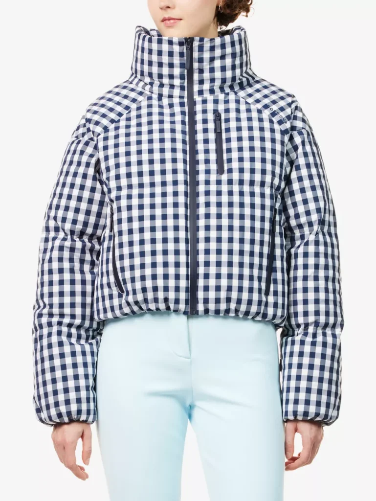 Model wearing the CORDOVA Cortina Vichy Shell Puffer Jacket in indigo gingham, featuring a cropped fit, padded high funnel neck, full zip closure, and quilted insulated construction, styled with light blue ski pants against a white background
