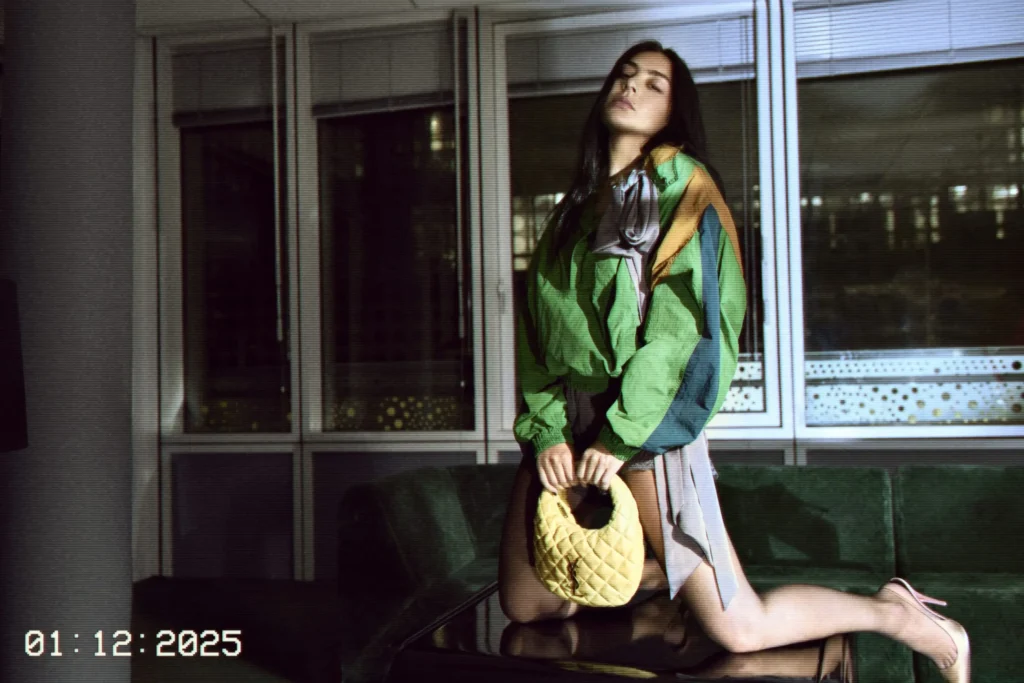 Charlie xcx on a glass table in a dimly lit room, wearing a multi-toned green and brown jacket paired with a short skirt and heels. She holds a small quilted yellow handbag with both hands while a timestamp reading 01:12:2025 appears in the lower left corner