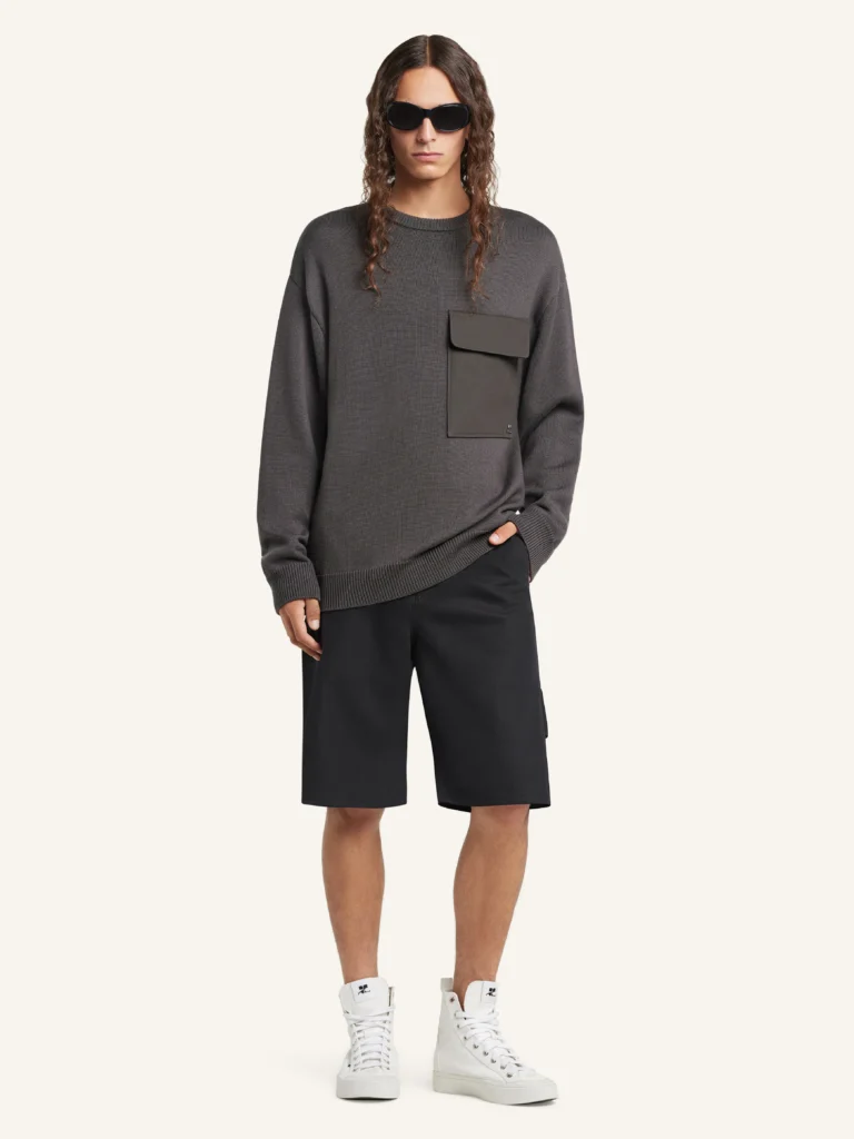 Model wearing the Courrèges Utility Wool Sweater in charcoal grey, styled with black tailored shorts and white high-top sneakers, showcasing the relaxed fit, structured knit, and functional chest pocket in a minimalist studio setting