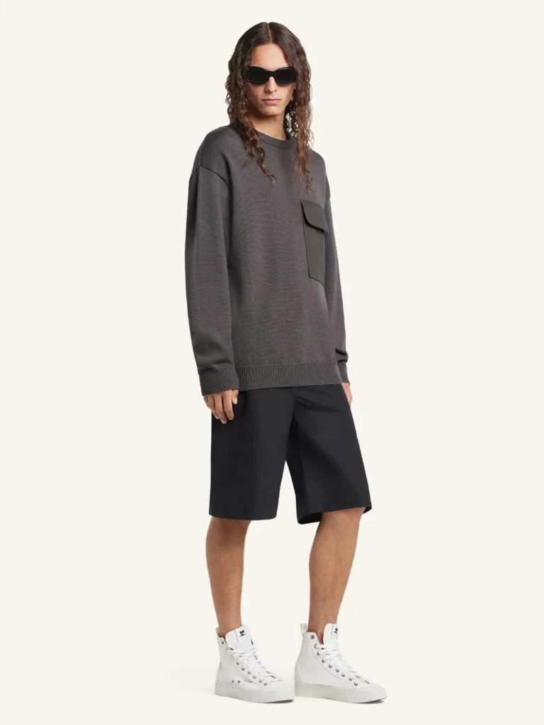 Side view of a model wearing the Courrèges Utility Wool Sweater in charcoal grey, highlighting the relaxed, architectural fit, clean crewneck, and utility-inspired chest pocket, styled with tailored black shorts and white high-top sneakers against a neutral studio backdrop