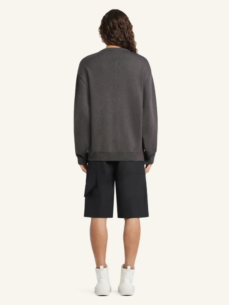 Back view of a model wearing the Courrèges Utility Wool Sweater in charcoal grey, showing the clean, uninterrupted knit back, relaxed silhouette, and ribbed hem, styled with black tailored shorts and white sneakers in a neutral studio setting