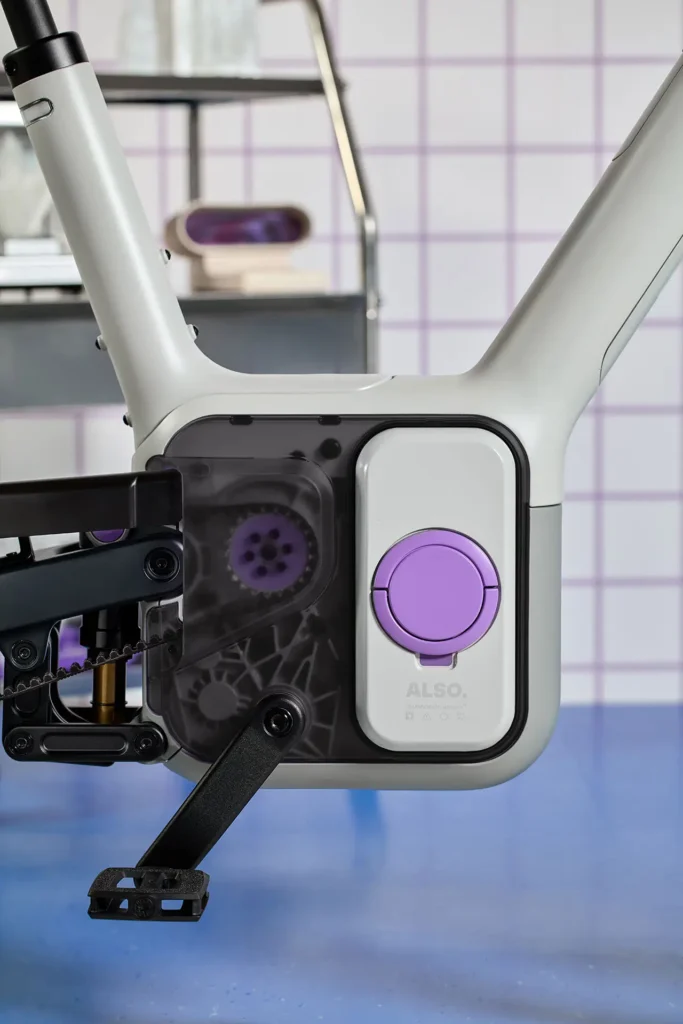 Close-up of the ALSO TM-B electric bike mid-drive and battery module, showing the translucent drivetrain housing, removable battery pack with purple accent, and clean modular frame construction that highlights its EV-inspired engineering design