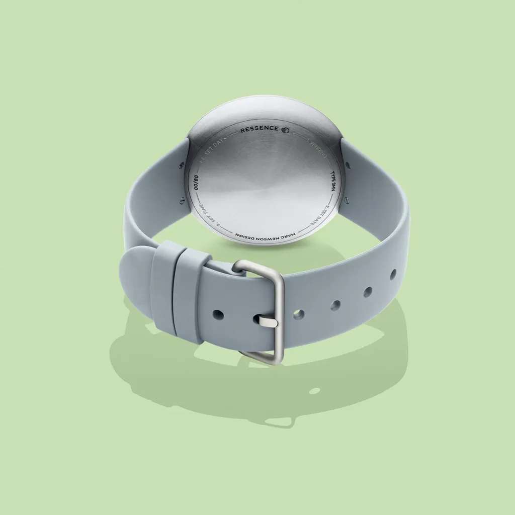 Rear view of the Ressence TYPE 3 MN watch by Marc Newson, showing the brushed titanium caseback with engraved markings, integrated lugs, and grey rubber strap with a minimalist metal buckle, photographed against a soft pastel green background