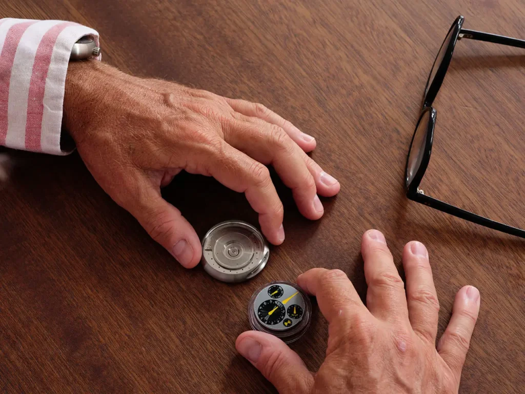 Close-up of hands disassembling the Ressence TYPE 3 MN watch on a wooden table, with the oil-filled dial module separated from the titanium caseback, highlighting the watch’s modular construction and mechanical design, alongside a pair of black eyeglasses