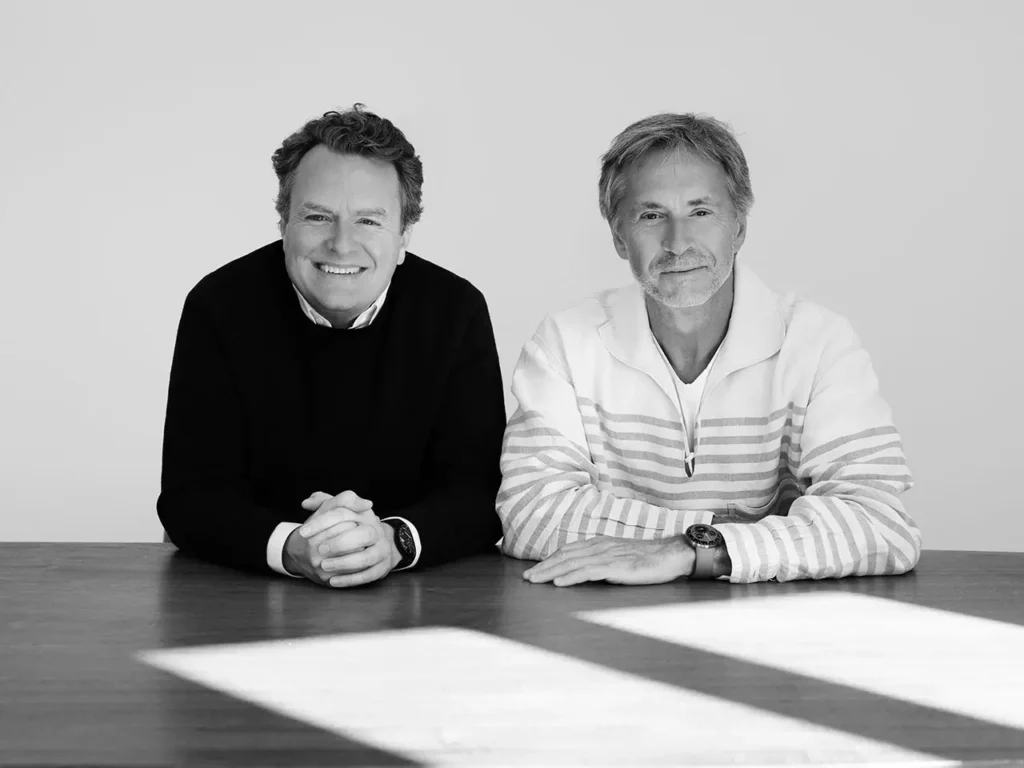 Black-and-white portrait of two men seated at a table, smiling toward the camera, representing Marc Newson and Benoît Mintiens of Ressence, photographed in a minimalist studio setting with soft natural light
