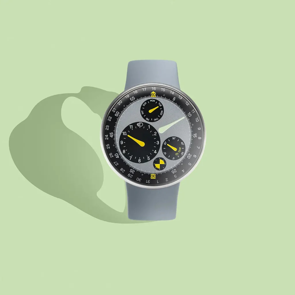Front-facing studio image of the Ressence TYPE 3 MN watch by Marc Newson, showing a floating oil-filled grey dial with rotating convex sub-dials, yellow accent hands, and a black minute track, set in a titanium case on a grey rubber strap against a soft pastel green background