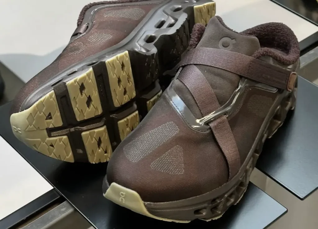 Brown performance sneakers displayed on a black platform at Paris Fashion Week, featuring translucent mesh uppers, fleece-lined collars, cross-strap closures, rugged segmented outsoles, and On branding on the toe, shown from an angled top-down view