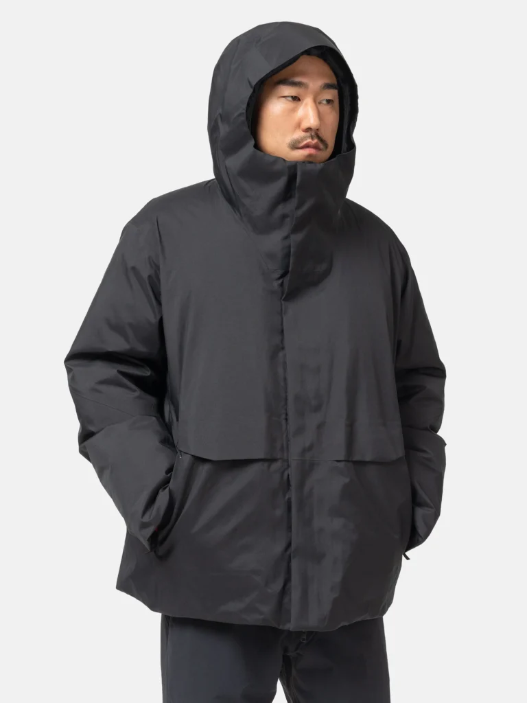 Model wearing the VEILANCE Sorin Down Jacket in black, shown in a three-quarter front view with the hood up, highlighting the jacket’s structured insulated silhouette, high protective collar, concealed front closure, matte technical fabric, and relaxed urban fit