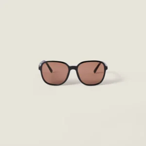 Front view of Miu Miu Regard sunglasses with a rounded black acetate frame, softly tinted brown lenses, and a clean, minimalist silhouette set against a light neutral background