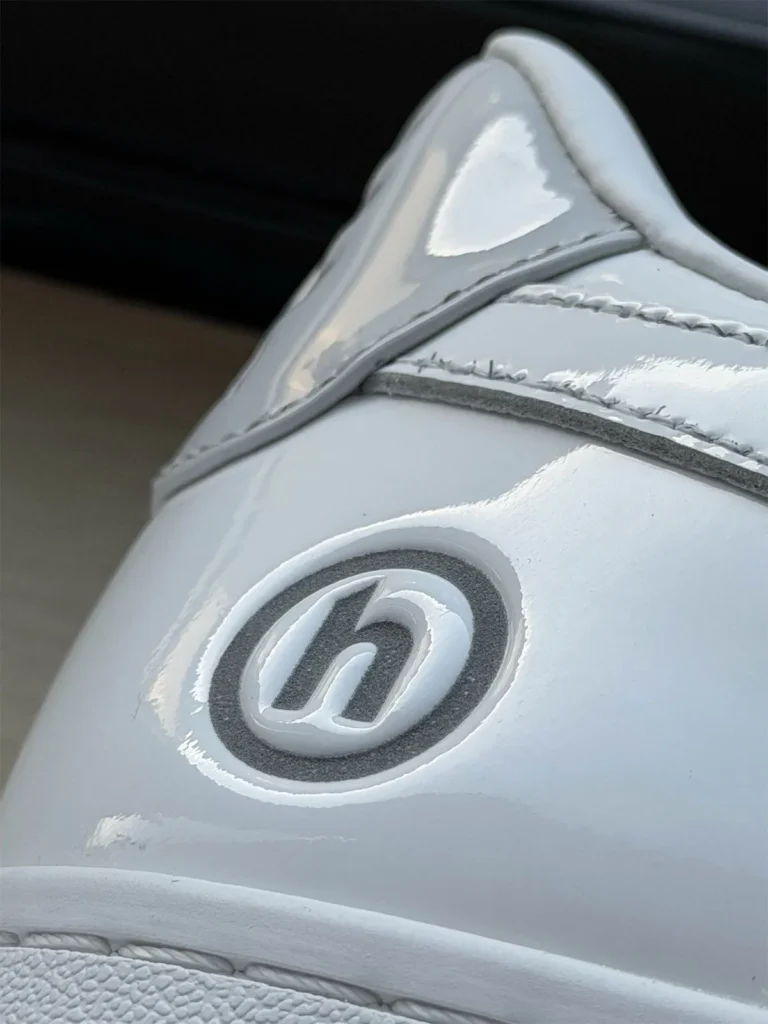 Close-up side view of a glossy white sneaker showing Hidden NY’s encircled “H” logo embossed on the heel panel, with reflective trim and stitching details that distinguish the BAPE Sta silhouette