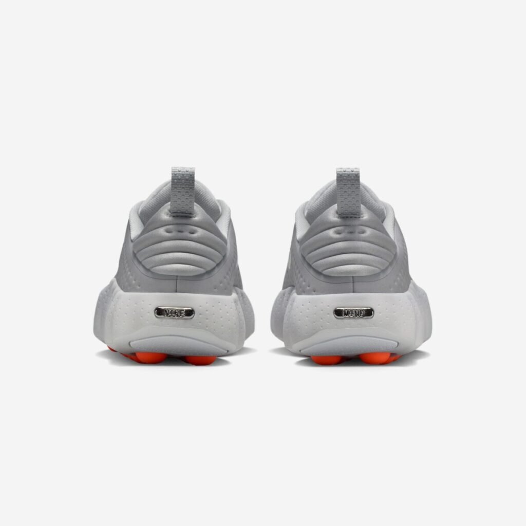 Rear view of the Nike Mind 002 “Light Smoke Grey” sneakers, showing the padded ribbed heel collars with pull tabs, perforated grey uppers, sculpted foam midsoles with chrome heel badges reading “Nike” and “Mind,” and the bright orange rounded outsole nodes peeking out beneath