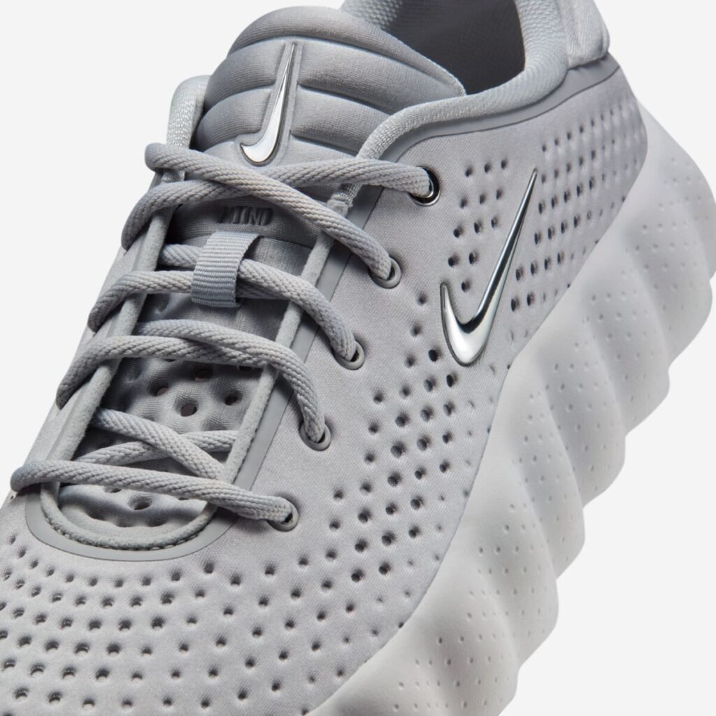 Close-up view of the Nike Mind 002 “Light Smoke Grey” sneaker, focusing on the perforated grey upper, tonal laces threaded through metal eyelets, padded tongue with subtle Nike branding, reflective chrome Swoosh, and the textured sculpted foam midsole along the side