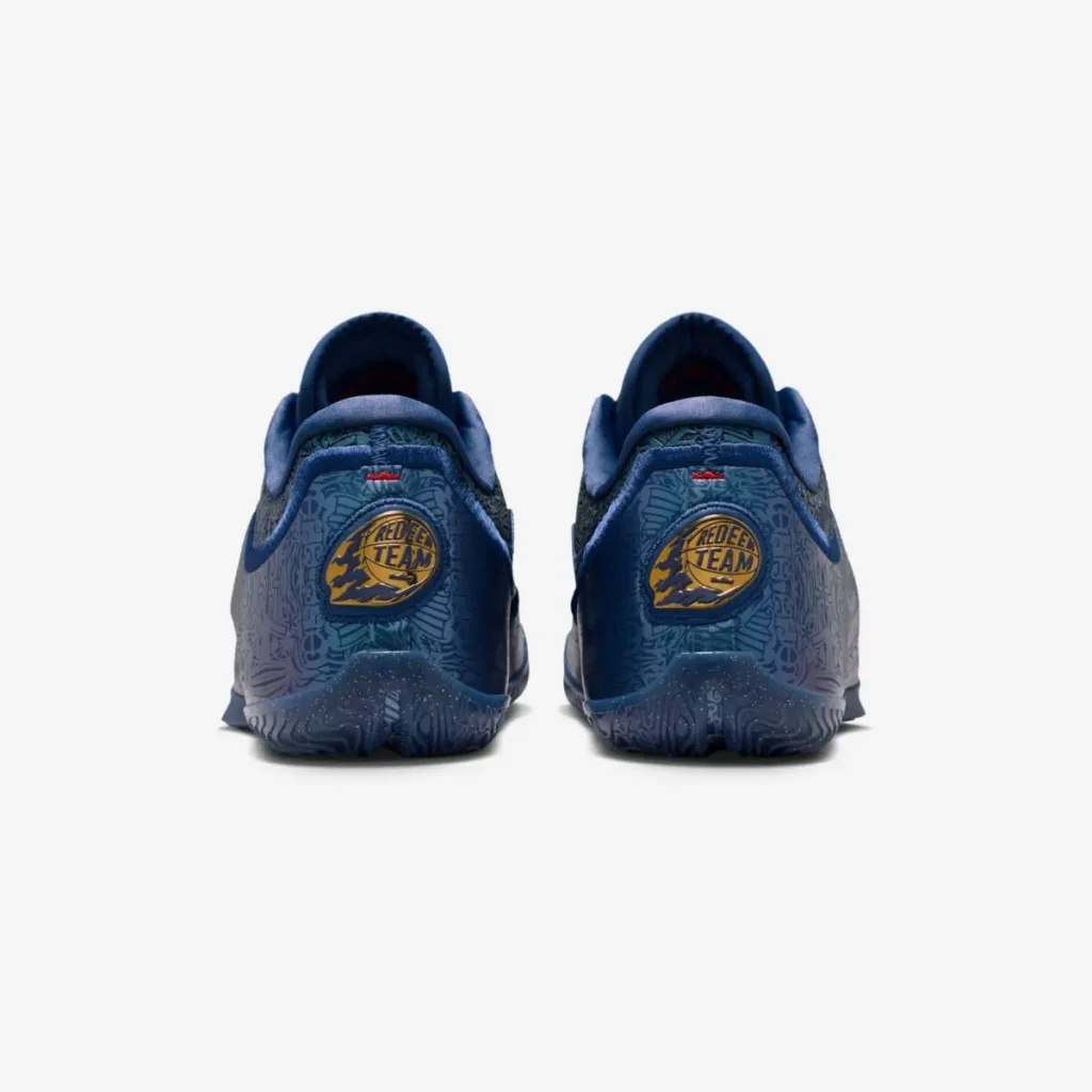 Rear view of the Nike LeBron 23 “Out For Redemption” sneakers in dark navy, showing embossed heel panels, circular gold-and-blue “Redeem Team” badges, padded collars, speckled outsole edges, and tonal graphic patterning on a white background