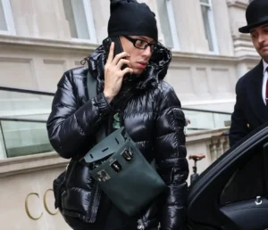 Central Cee wearing glossy black puffer jacket and black head covering talks on a phone while stepping out of a car, carrying a structured dark green crossbody bag with metal buckle details in an urban street setting