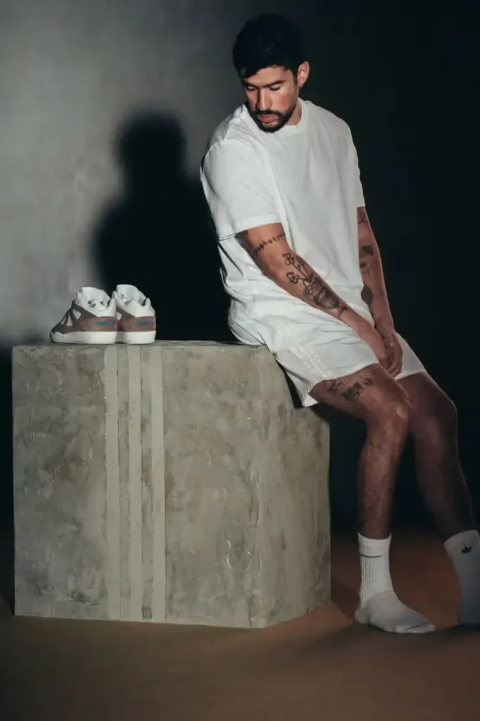 Bad Bunny crouches on a concrete plinth wearing white socks while his adidas BadBo 1.0 sneakers sit beside him in a dramatic studio-lit setting