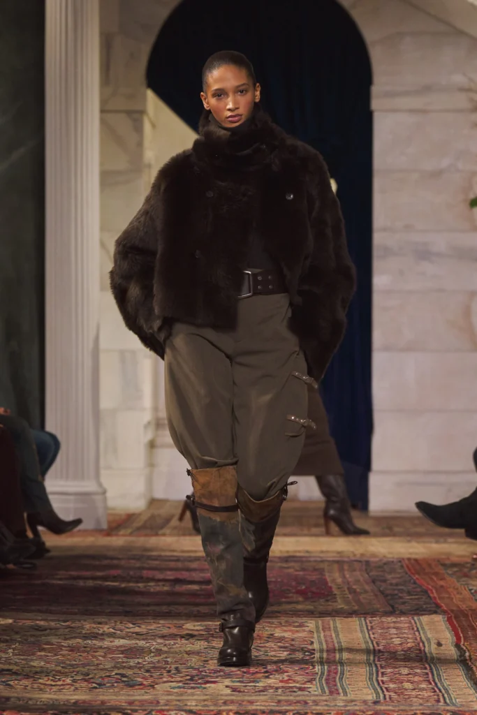Model walking the runway at the Ralph Lauren Fall/Winter 2026 show wearing a dark cropped fur jacket, high-waisted tailored trousers, and weathered knee-high riding boots. The look is styled with a wide belt and presented against a classical interior with marble columns, dark drapery, and a vintage patterned carpet