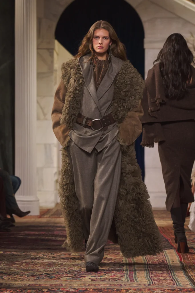 Model walks the runway at the Ralph Lauren Fall/Winter 2026 show wearing a layered grey tailored suit beneath a long shearling-trimmed coat, cinched with a wide leather belt. The look blends soft textures and structured tailoring on a richly patterned runway set inside a classical space with marble columns and dark drapery