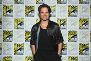 Sebastian Stan smiles while posing at San Diego Comic-Con in front of a repeating Comic-Con International logo wall, wearing a black shirt, textured short-sleeve jacket, gold chain necklace, and wristwatch with his hands in his pockets