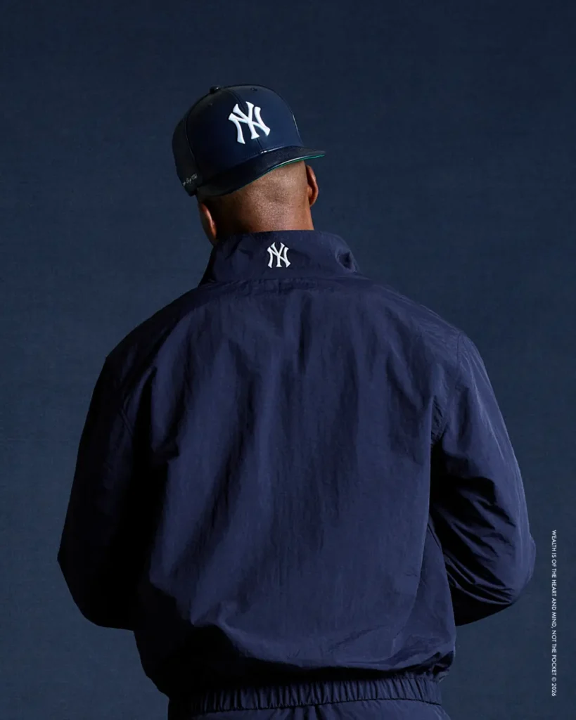 Back view of a model wearing a navy New York Yankees cap paired with a matching navy windbreaker jacket, both featuring subtle white NY logos, styled against a dark blue studio backdrop for a minimal, elevated streetwear look